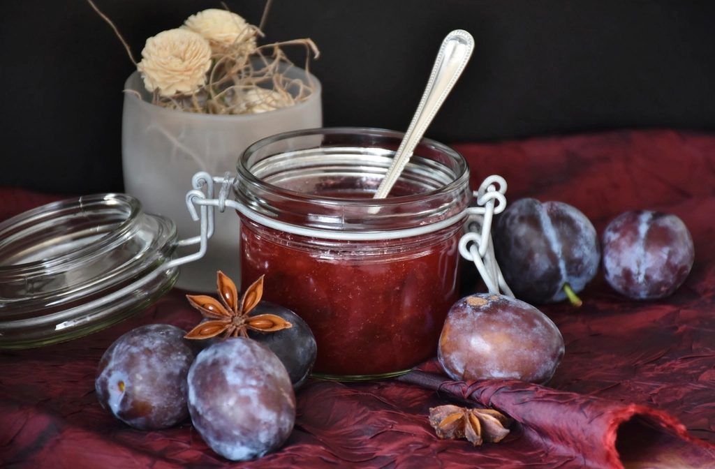 Open jar of dark preserves with a spoon and fresh plums